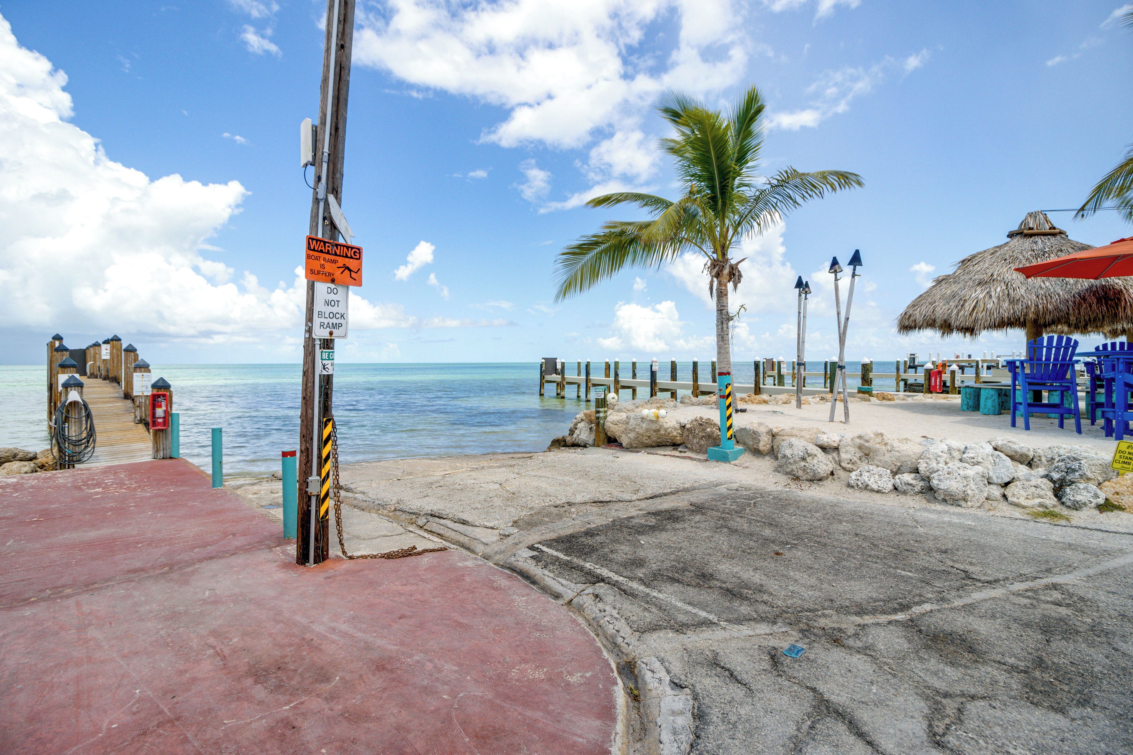 A beach scene with a wooden pier, a thatched umbrella, and blue chairs.