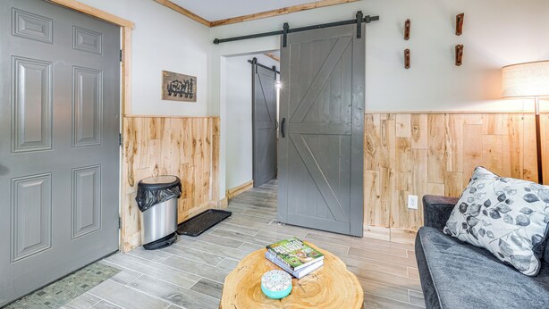 A room with a grey door, a sliding barn-style door, a wooden wall panel, a grey sofa with a patterned pillow, and a small table with a book and a decorative item.