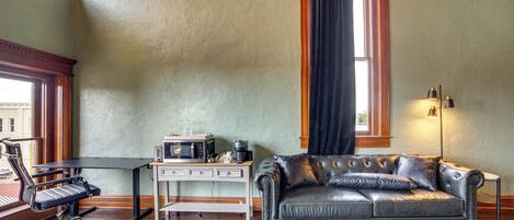 A room with a leather sofa, a desk with a computer and coffee maker, and a window with curtains.
