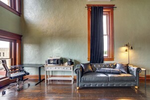 Una habitación con sofá de cuero, escritorio con computadora y cafetera, y ventana con cortinas.