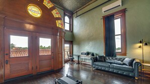 Una habitación amplia con suelos de madera, un sofá de cuero, un escritorio con ordenador y ventanas grandes con cortinas.