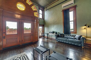 Una habitación amplia con suelos de madera, un sofá de cuero, un escritorio con ordenador y ventanas grandes con cortinas.