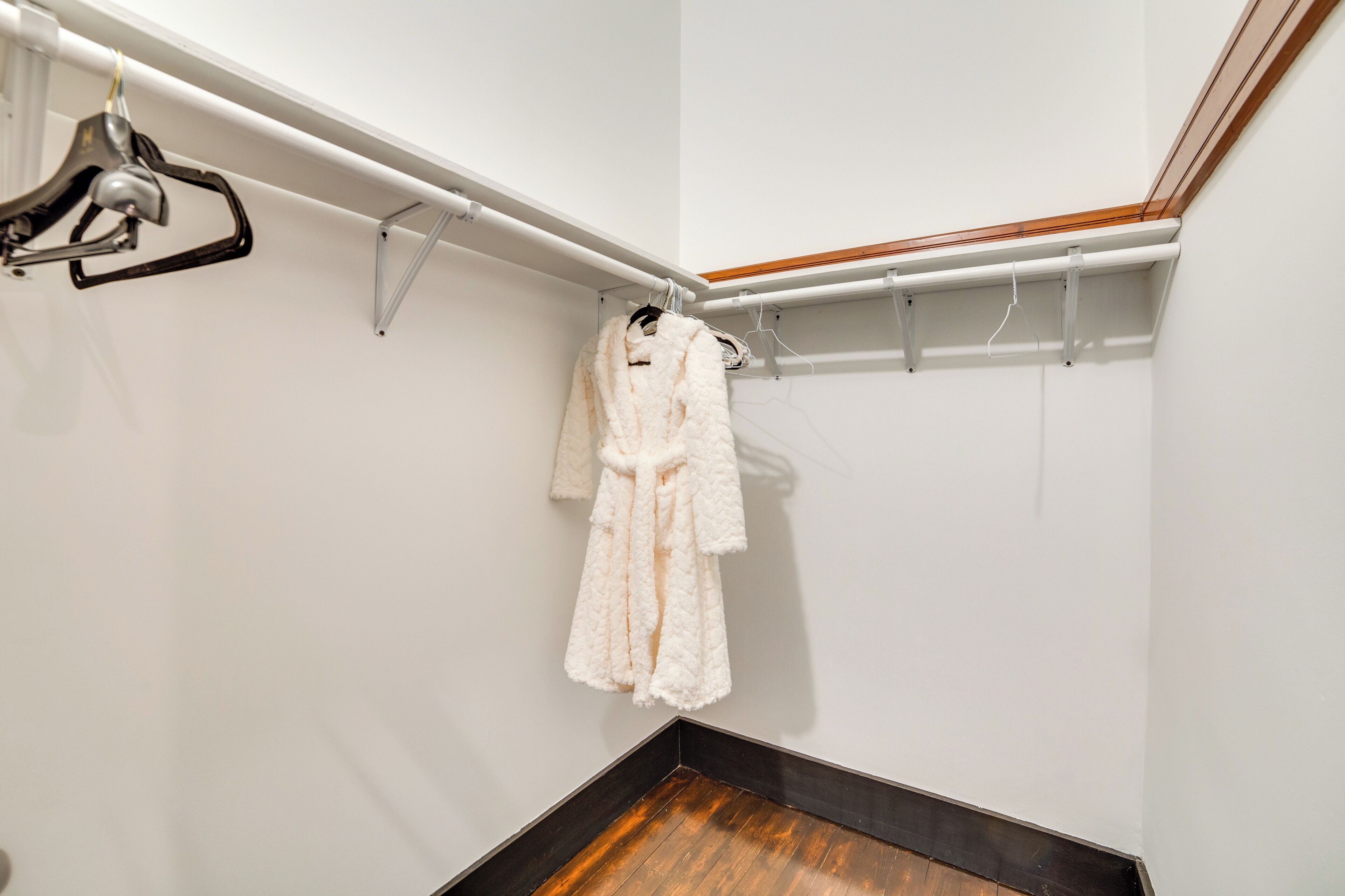 A white robe hanging on a metal rack in a corner of a room with white walls and a wooden floor.