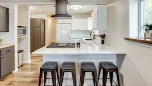 A modern kitchen with a central island, bar stools, stainless steel appliances, and a window with a view. - Baldwin Lakefront Home: Dock, Fire Pit & Trails! (Baldwin)
