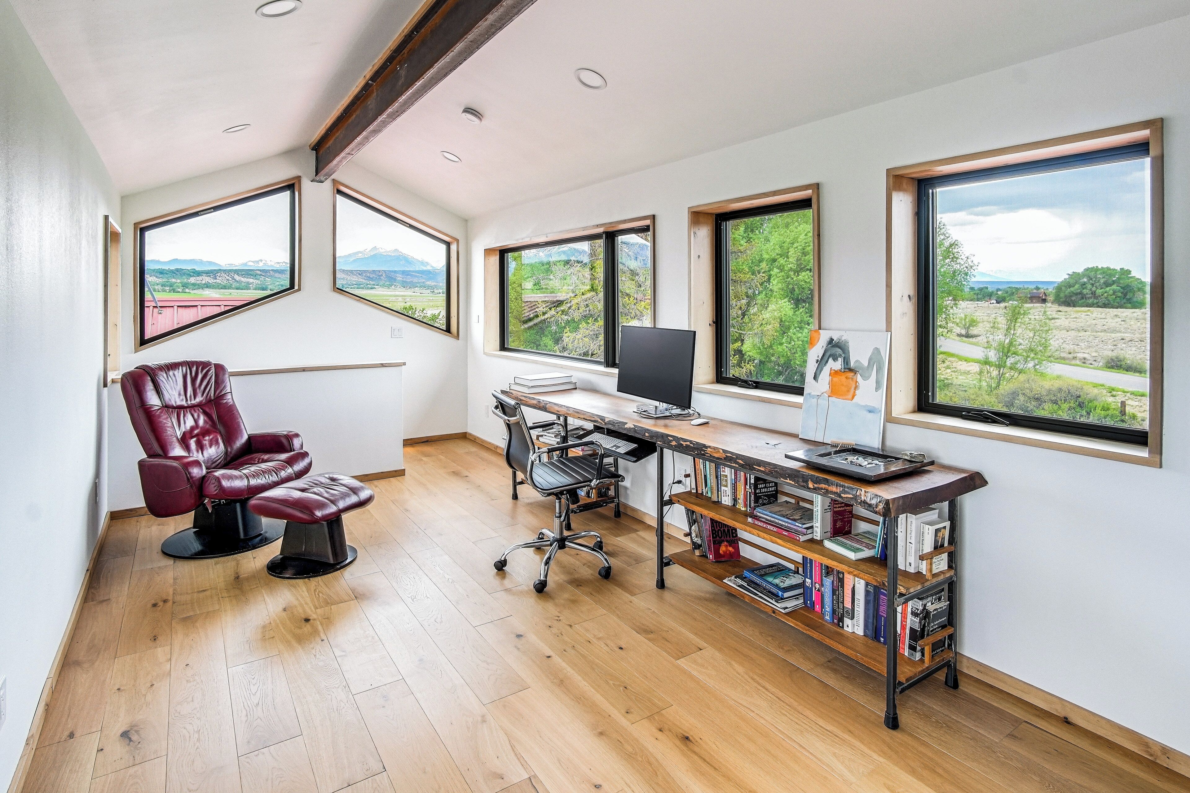 A modern office space with a leather chair, a desk with a computer, and a bookshelf. Large windows provide views of the outdoors.