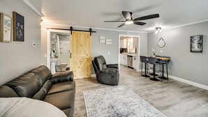 Una sala moderna con un sofá de cuero, un sillón reclinable negro, una puerta de madera, una zona de cocina y una mesa de comedor con taburetes de bar.