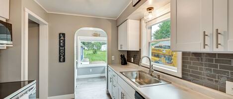 A modern kitchen with white cabinets, a stainless steel sink, and a dishwasher. There is a window with a view of the outside, and a sign on the wall that reads "LAKE HOUSE."