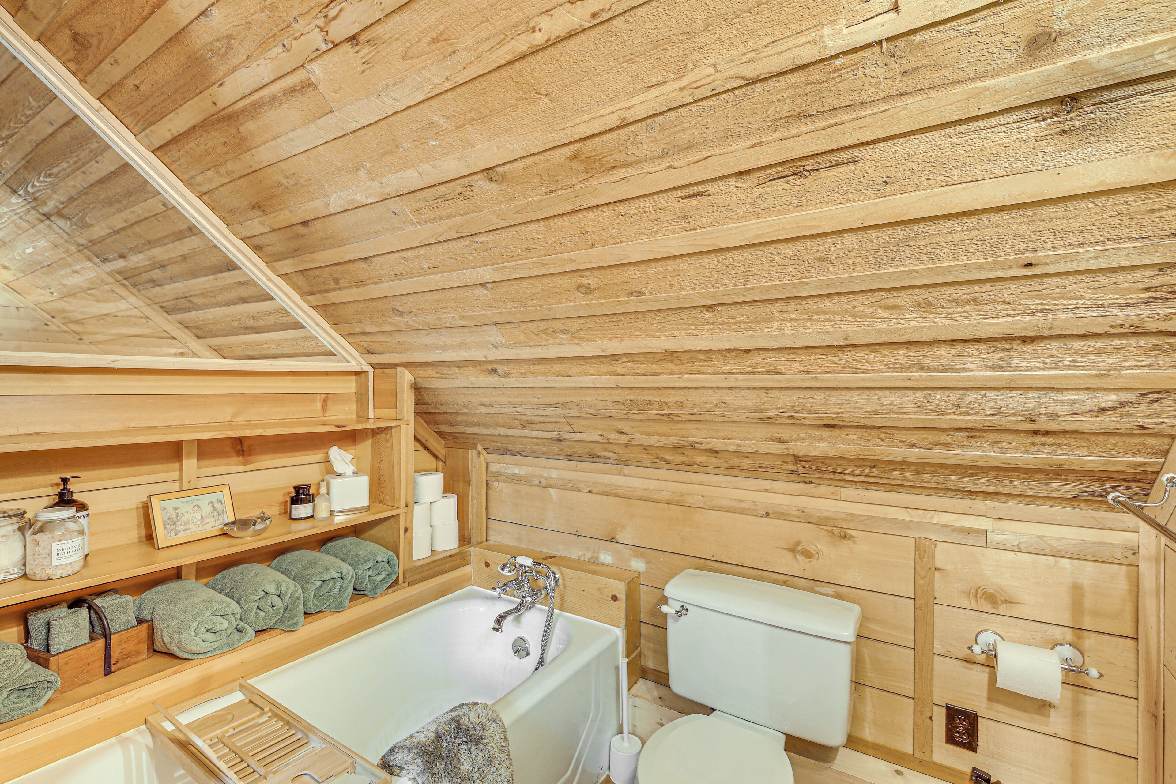 A wooden cabin bathroom with a bathtub, toilet, shelves with toiletries, and towels.