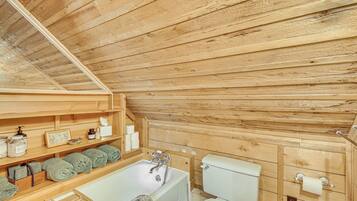 A wooden cabin bathroom with a bathtub, toilet, shelves with toiletries, and towels.