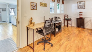 Un bureau à domicile comprenant un bureau, un fauteuil de bureau et une table à manger avec des chaises. Les murs sont ornés de tableaux encadrés et il y a une fenêtre avec des rideaux.