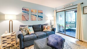 A living room with a dark blue sofa, two lamps, a coffee table, and a rug. There are three wall art pieces depicting beach scenes. A sliding glass door leads to an outdoor seating area. - Heated Pool & Hot Tub! Walk to Beach & Apache Pier (Myrtle Beach)