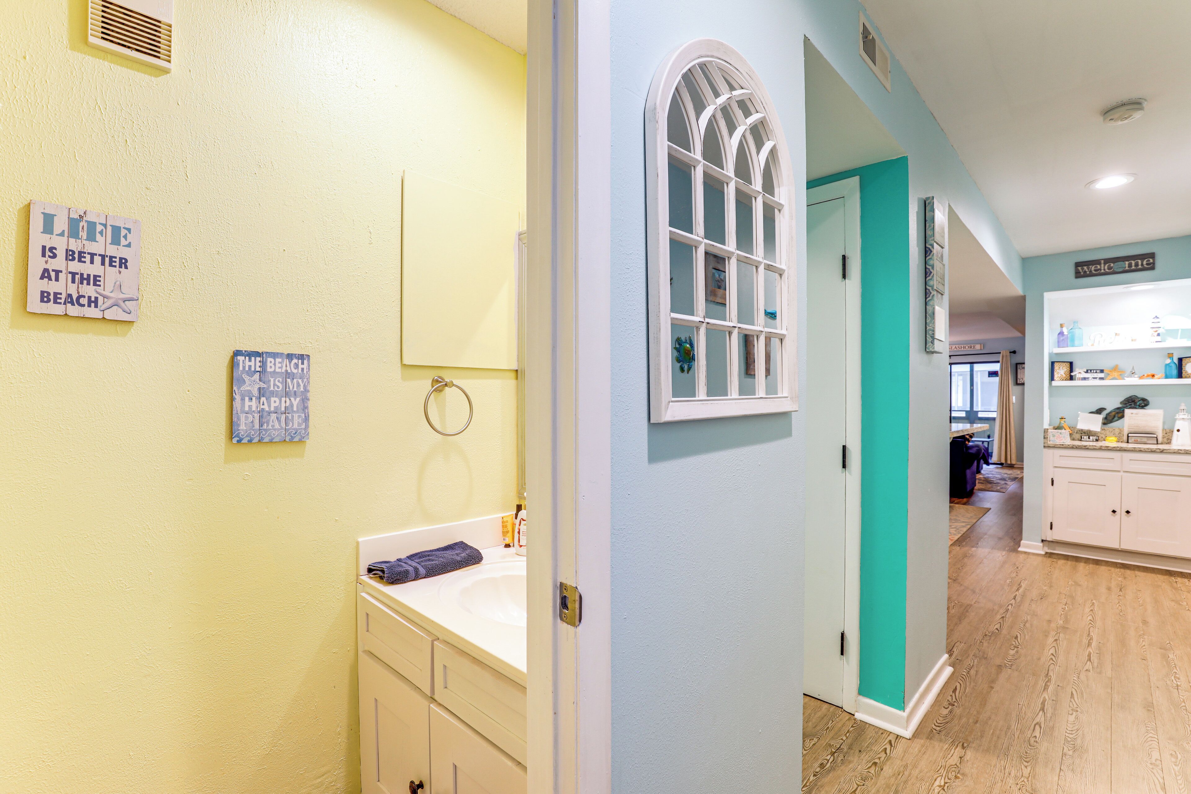 A hallway with a bathroom on the left, featuring a sink, mirror, and towel rack. The walls are painted in light yellow and teal, and there are signs on the wall.