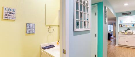 A hallway with a bathroom on the left, featuring a sink, mirror, and towel rack. The walls are painted in light yellow and teal, and there are signs on the wall.