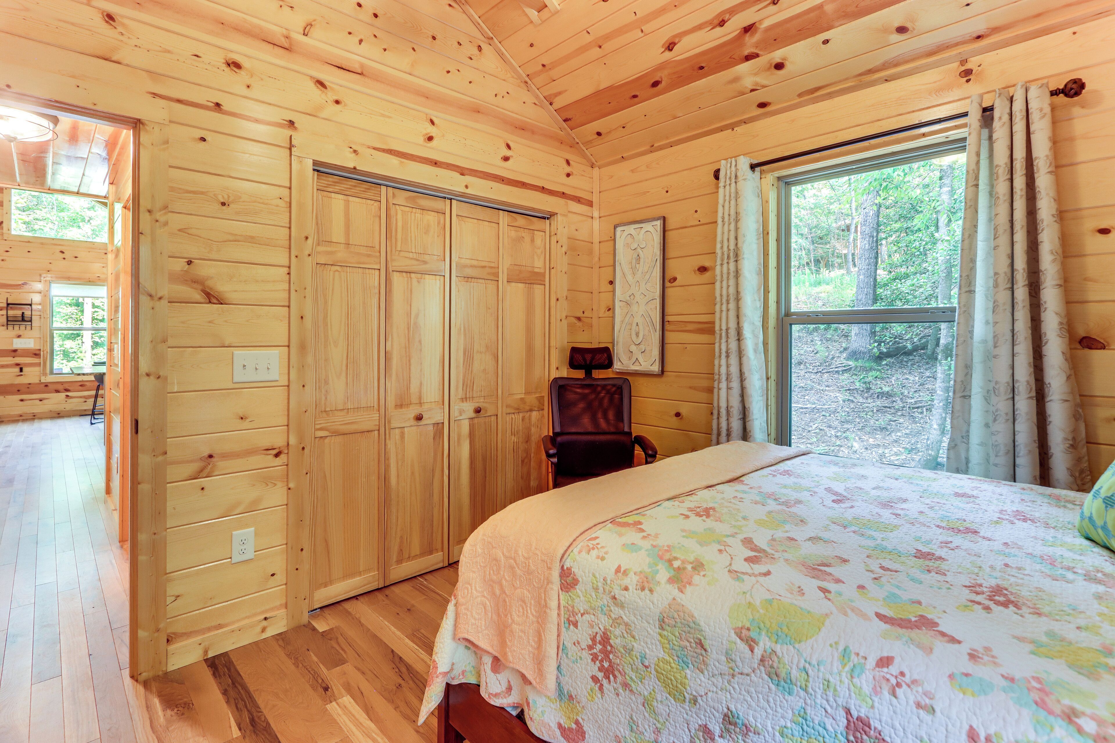 A wooden cabin bedroom with a bed, a chair, a wardrobe, and a window with curtains.