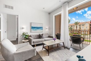 A modern living room with a sofa, armchair, and coffee table, a painting on the wall, and a view of a residential area through a balcony.