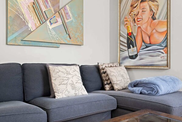 A living room with a grey sectional sofa, patterned cushions, a glass coffee table, and two artworks on the wall.