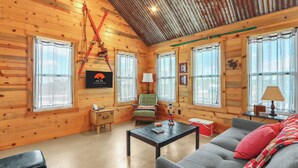 Ein gemütliches Blockhaus-Interieur mit Holzwänden, einer hohen Decke und einem Kamin. Ein grüner Sessel, ein graues Sofa und ein kleiner Tisch mit Büchern sind ebenfalls vorhanden.