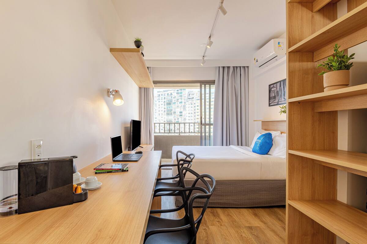 Ein modernes Hotelzimmer mit einem Holztisch, einem Bett mit blauem Kopfkissen, einem Laptop und einem Fenster mit Stadtblick.
