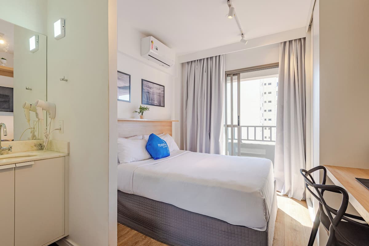 Ein Hotelzimmer mit Bett, Schreibtisch, Stuhl, einem fenster mit Vorhängen, einem Spiegel und einer Klimaanlage.