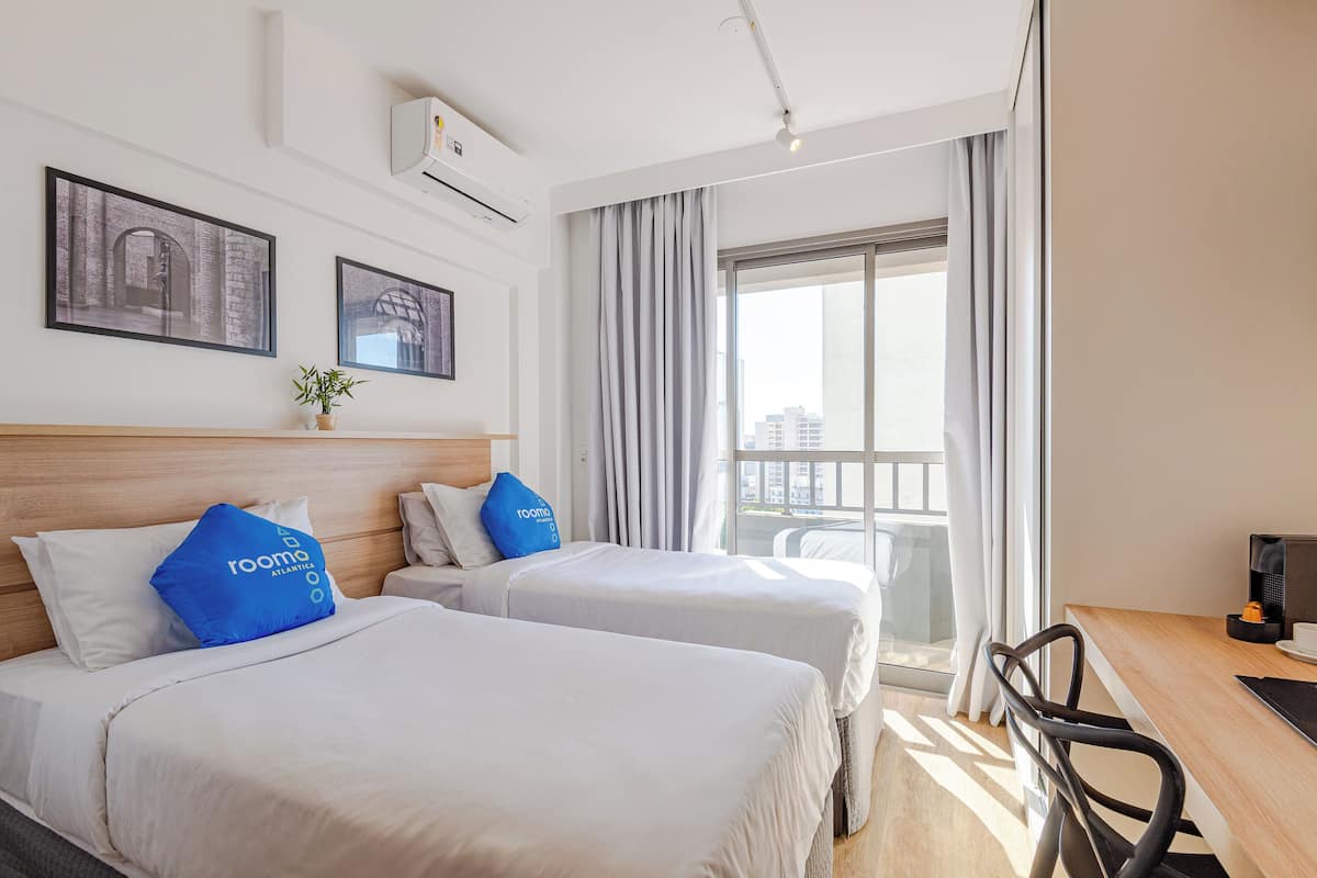 Ein Hotelzimmer mit zwei Betten, einem Schreibtisch, einem Stuhl und einem großen Fenster mit Vorhängen.