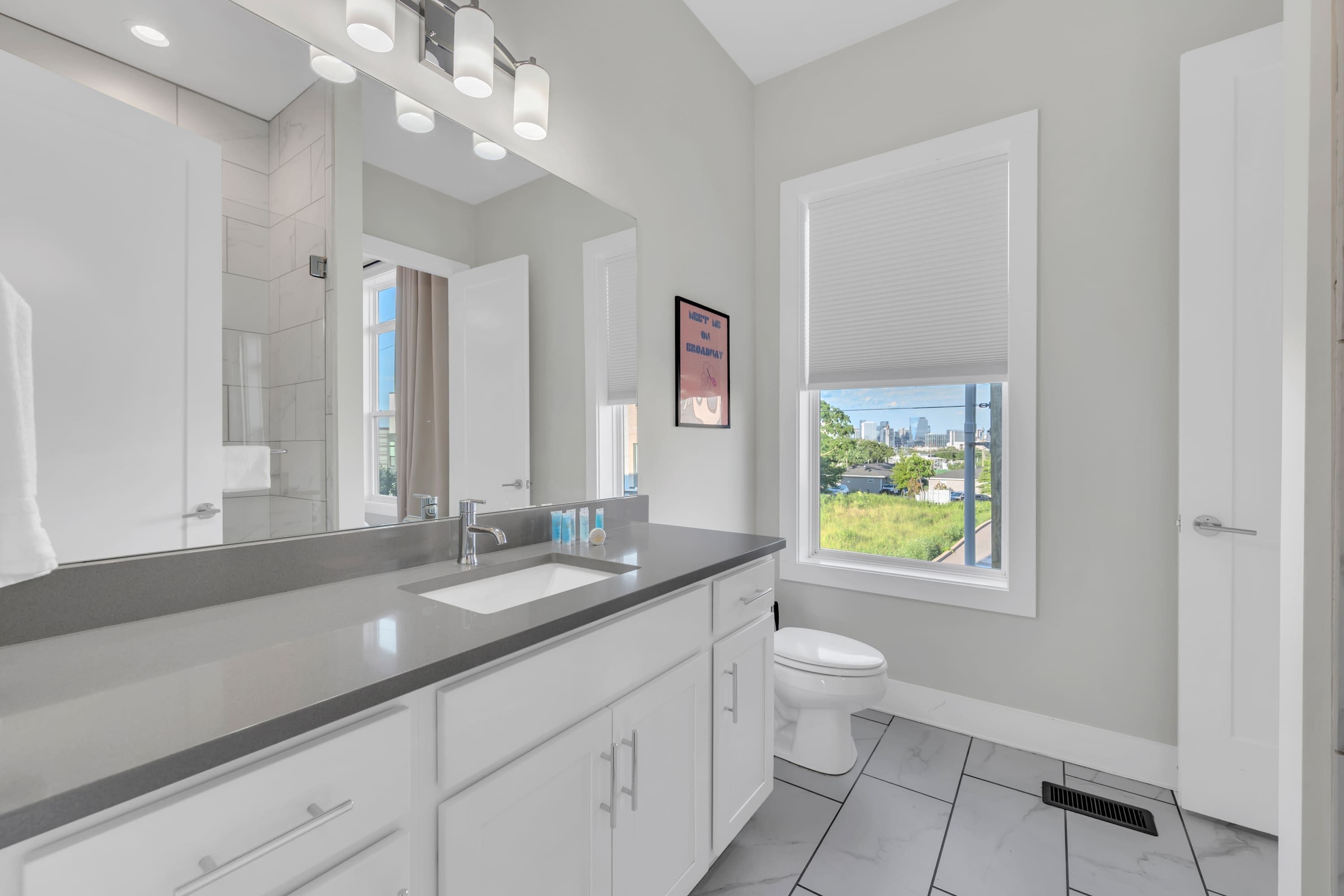 A modern bathroom with a large mirror, a sink, and a toilet. There is a window with a view of the city.