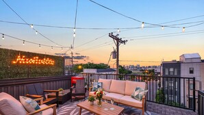 Une terrasse sur le toit, avec un canapé, des chaises, une table avec des boissons, des guirlandes lumineuses et un panneau néon.