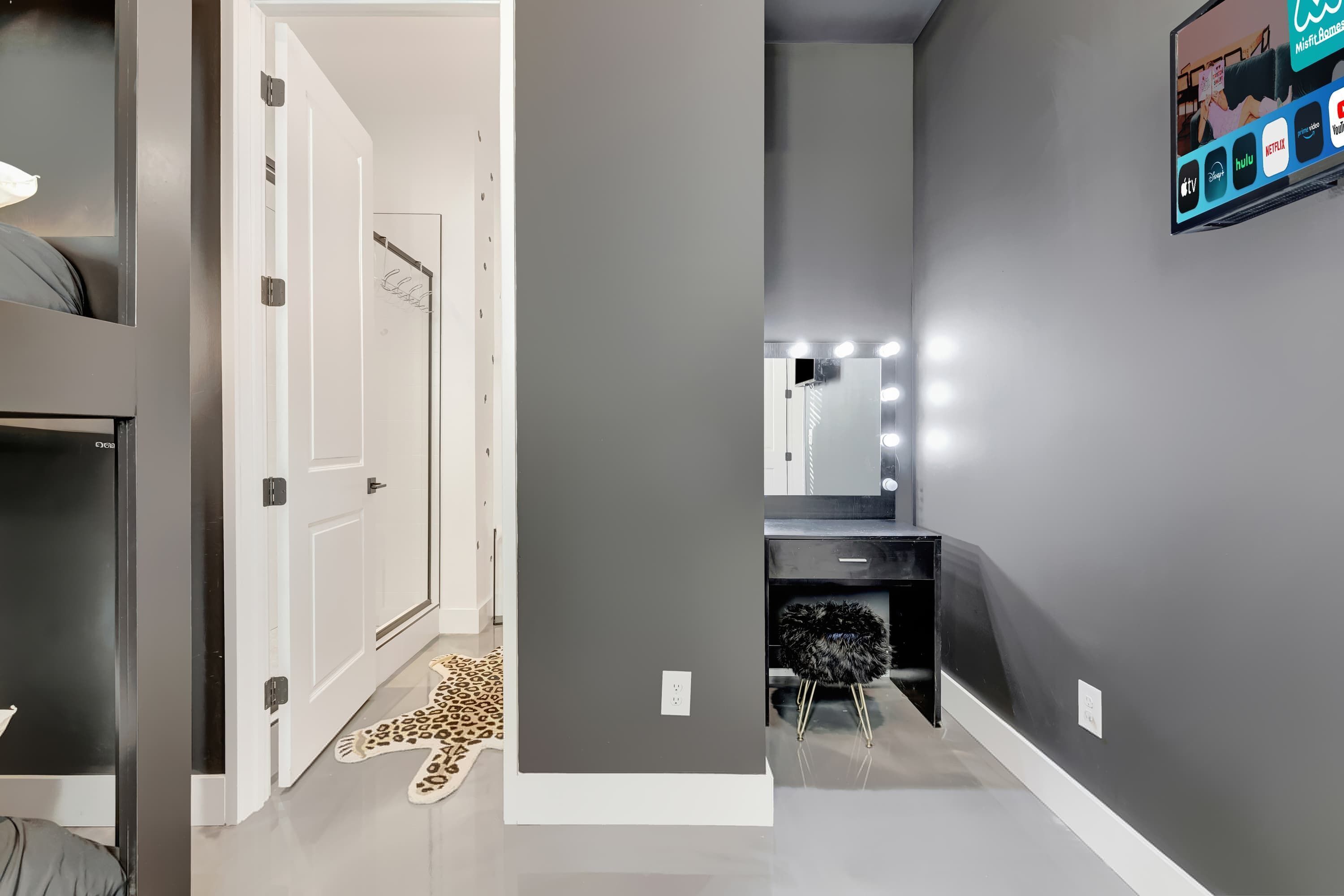 Un couloir avec un mur gris, une porte blanche, un tapis à motifs léopard, une chaise noire et une coiffeuse avec un miroir et des lampes.