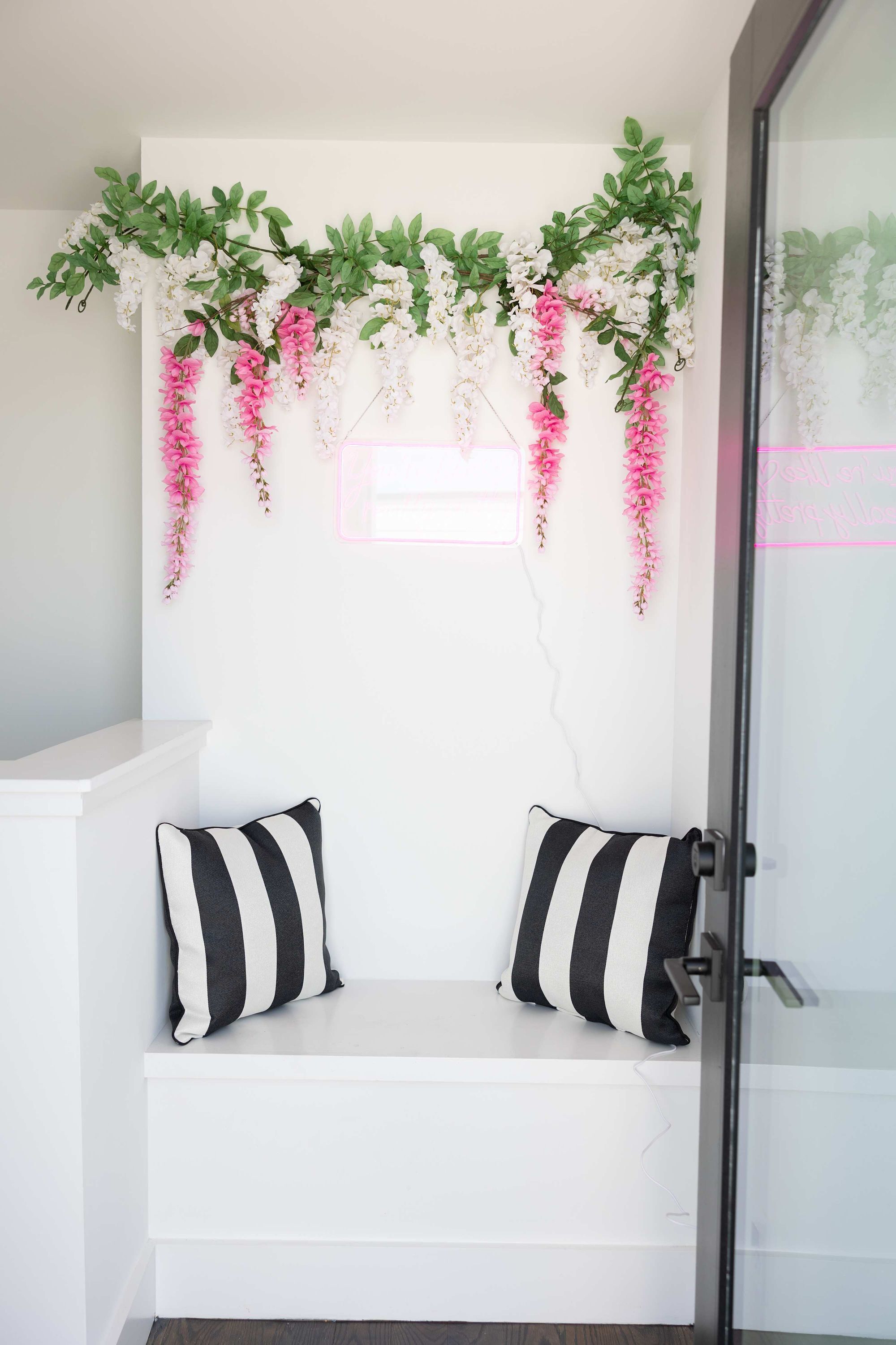 A white bench with black and white striped pillows against a wall with hanging greenery and flowers.