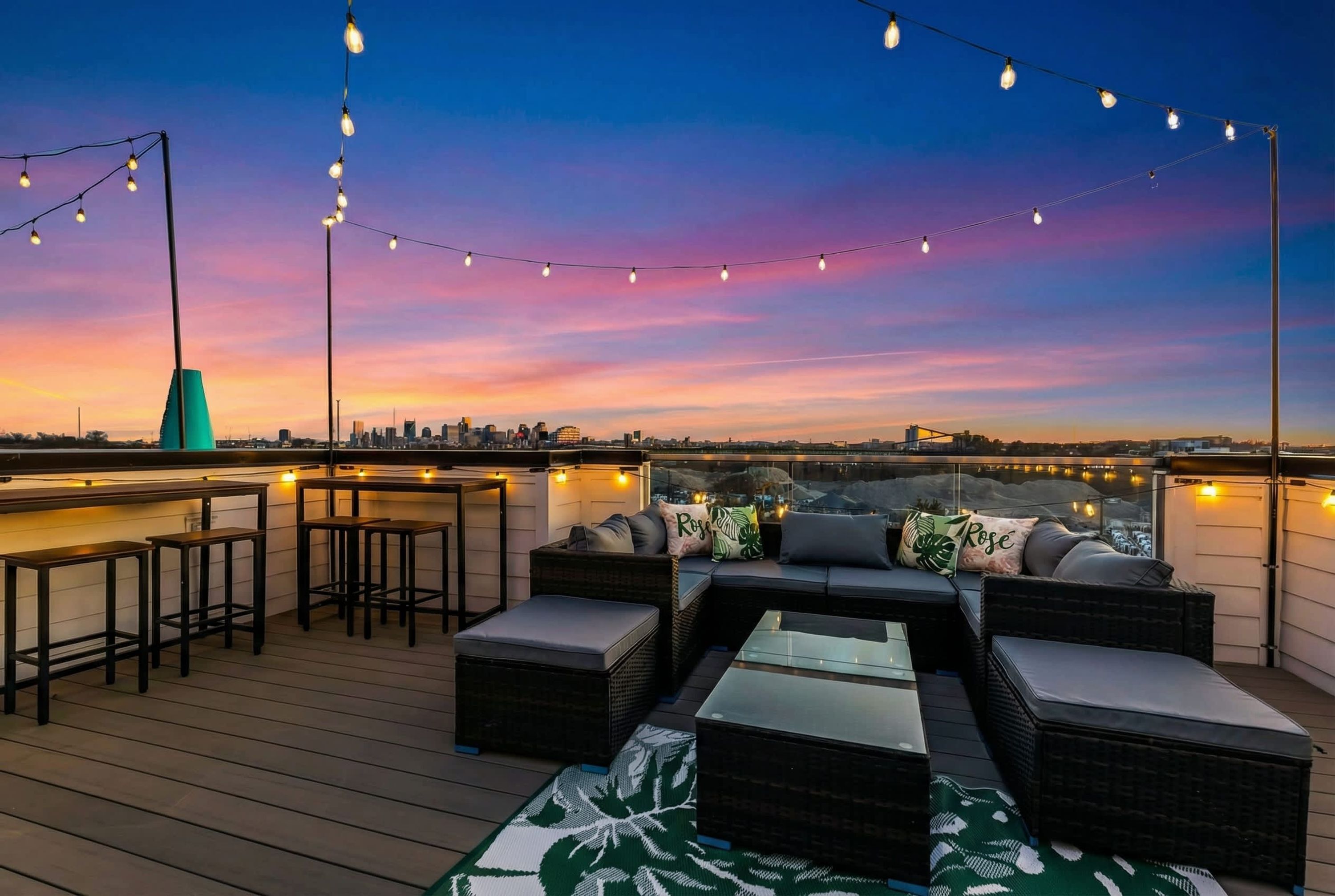 A rooftop bar with wicker furniture, string lights, and a city view at sunset.