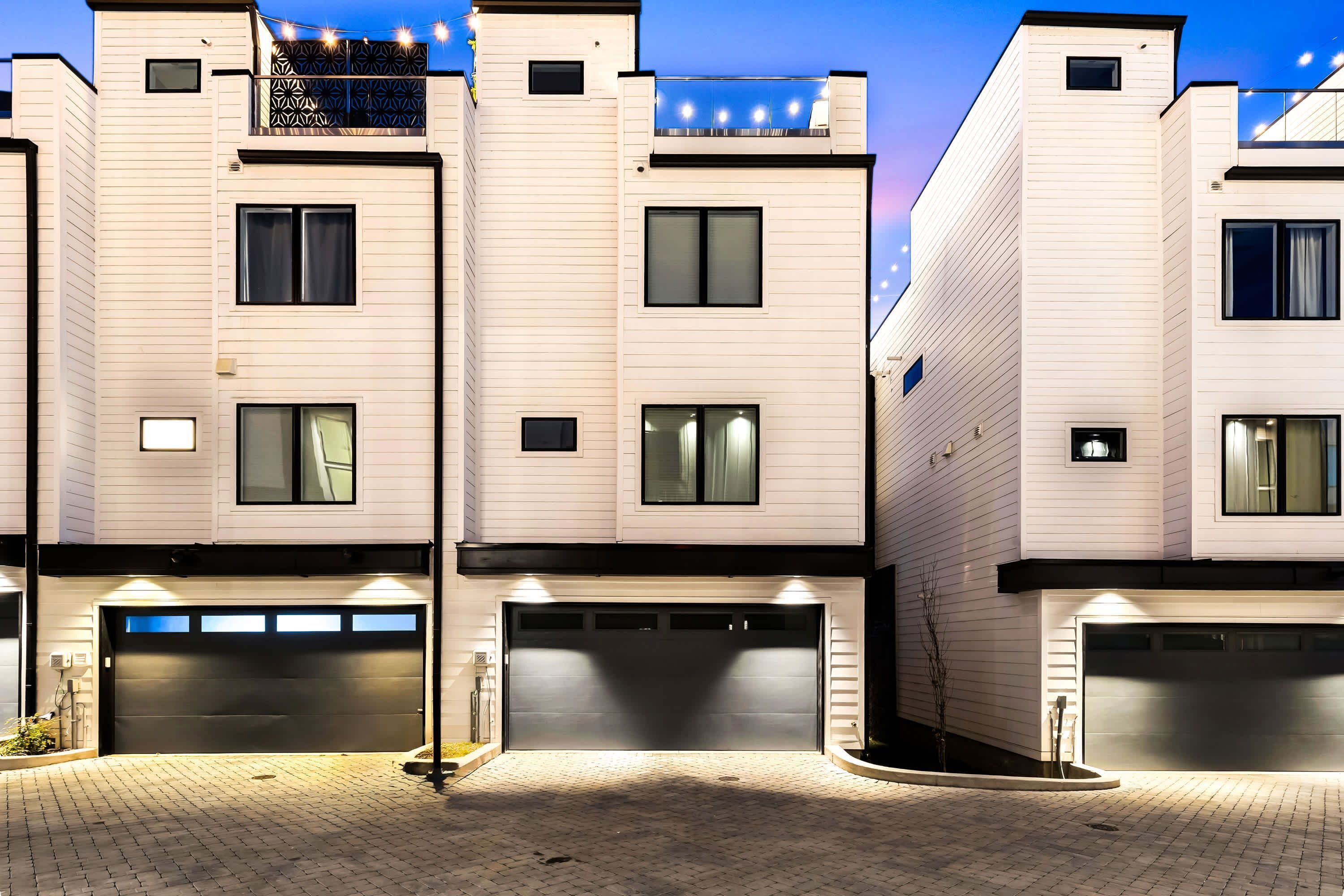 A modern multi-story residential building with a brick driveway and illuminated garage doors.