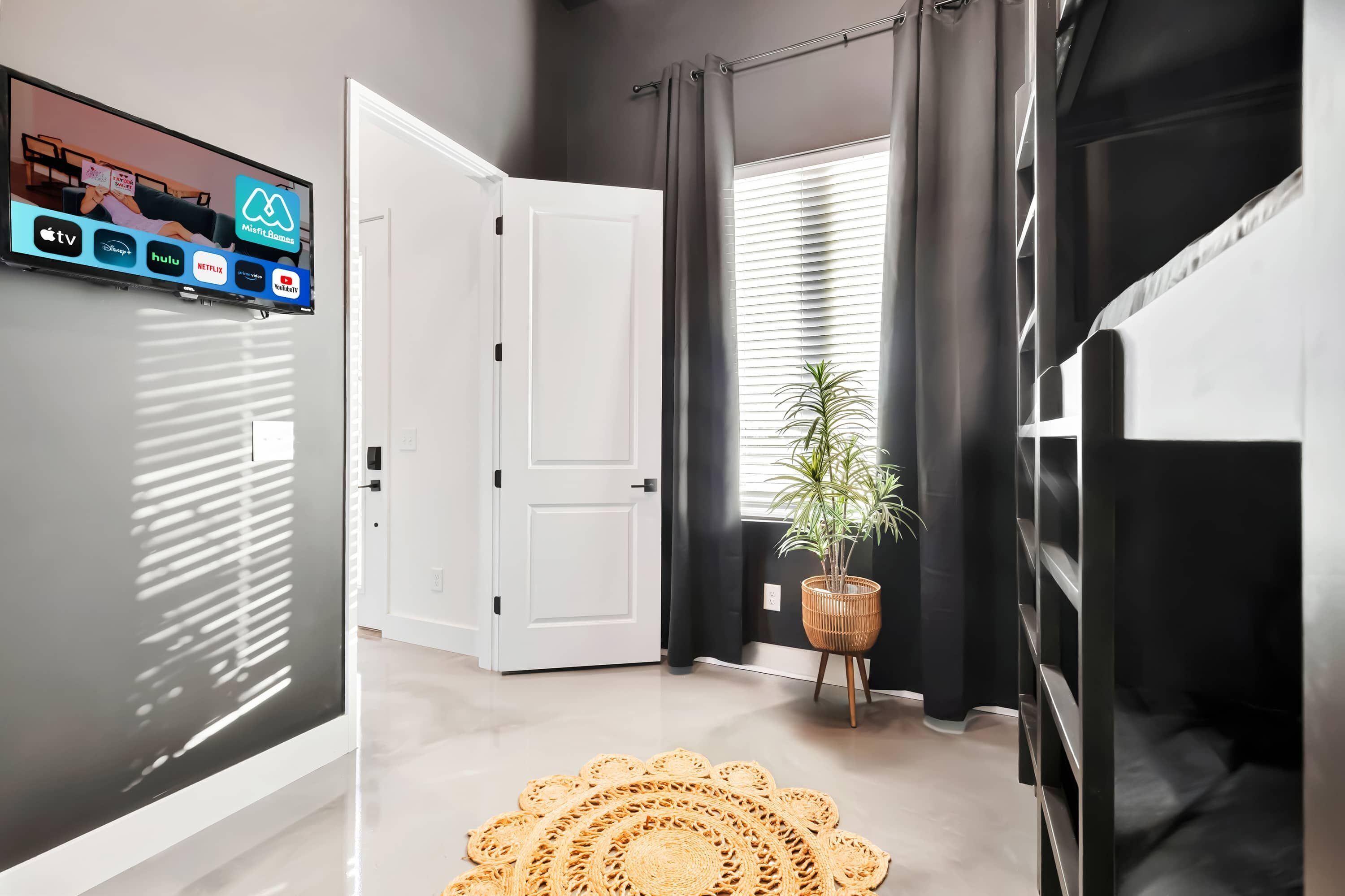 Una habitación de hotel moderna con una televisión de pantalla plana montada en la pared, una puerta blanca, una ventana con persianas, una planta en maceta y una cama litera.