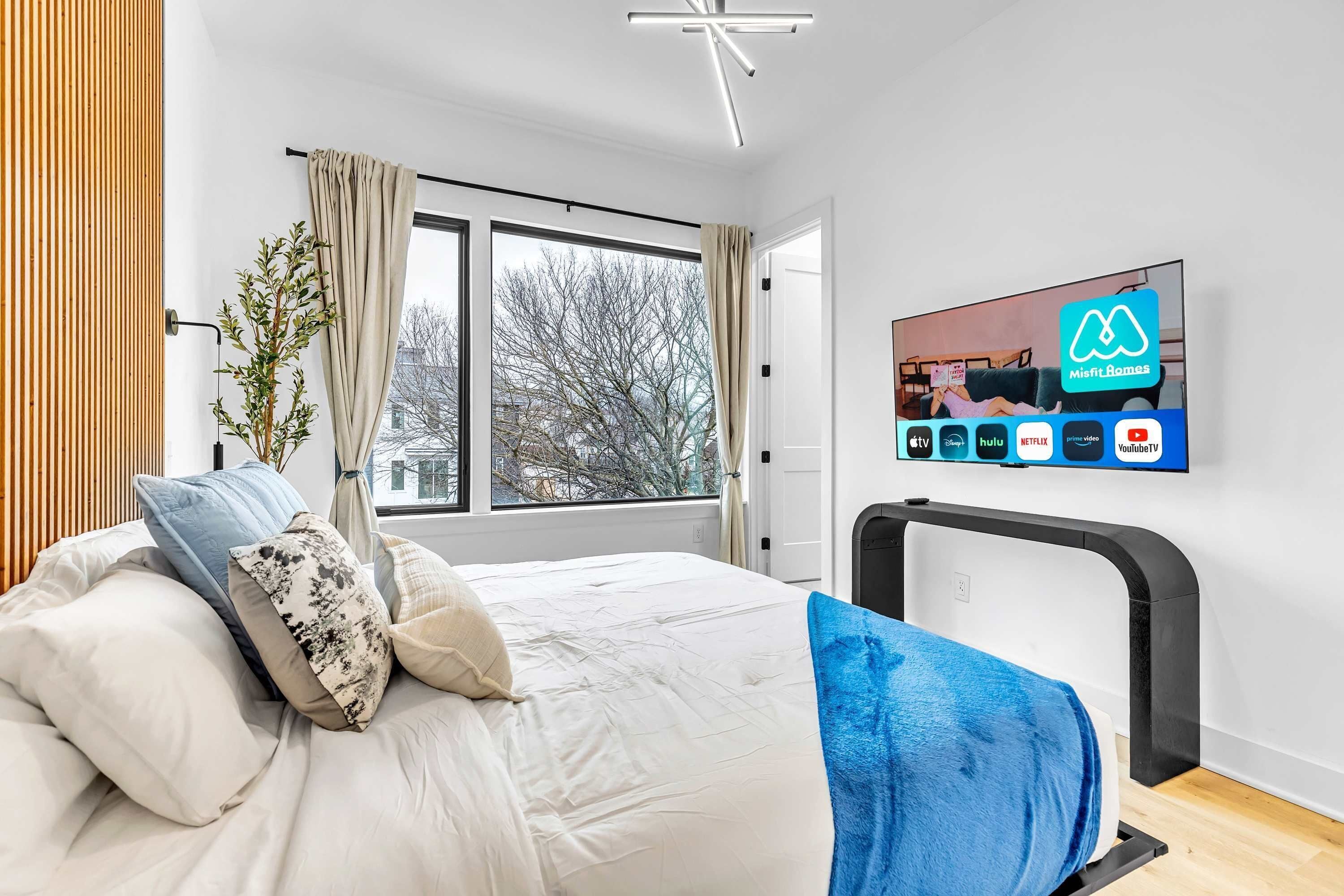 A modern bedroom with a bed, a TV displaying streaming services, and a window with curtains.