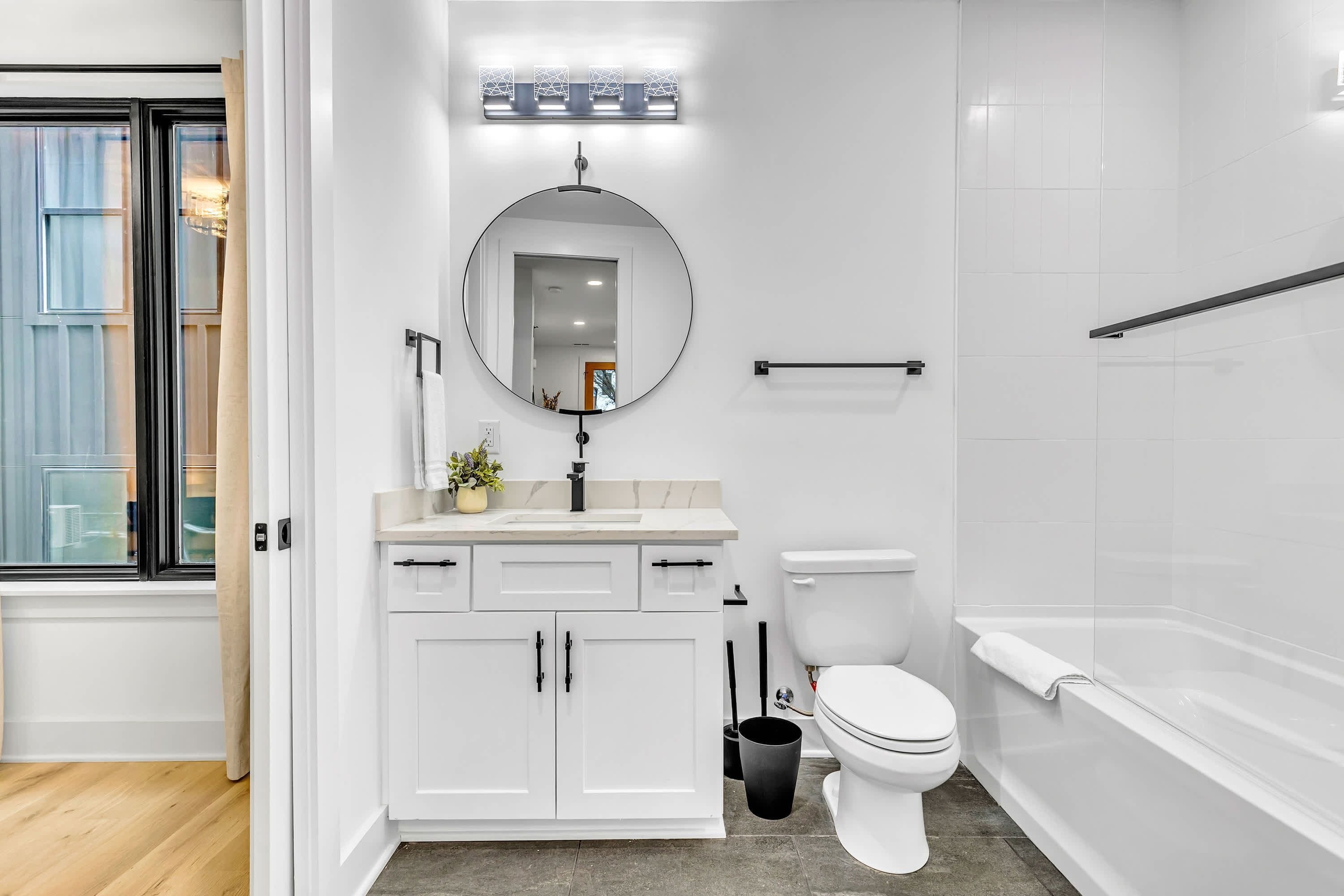 A modern bathroom with a white vanity, round mirror, toilet, and bathtub.
