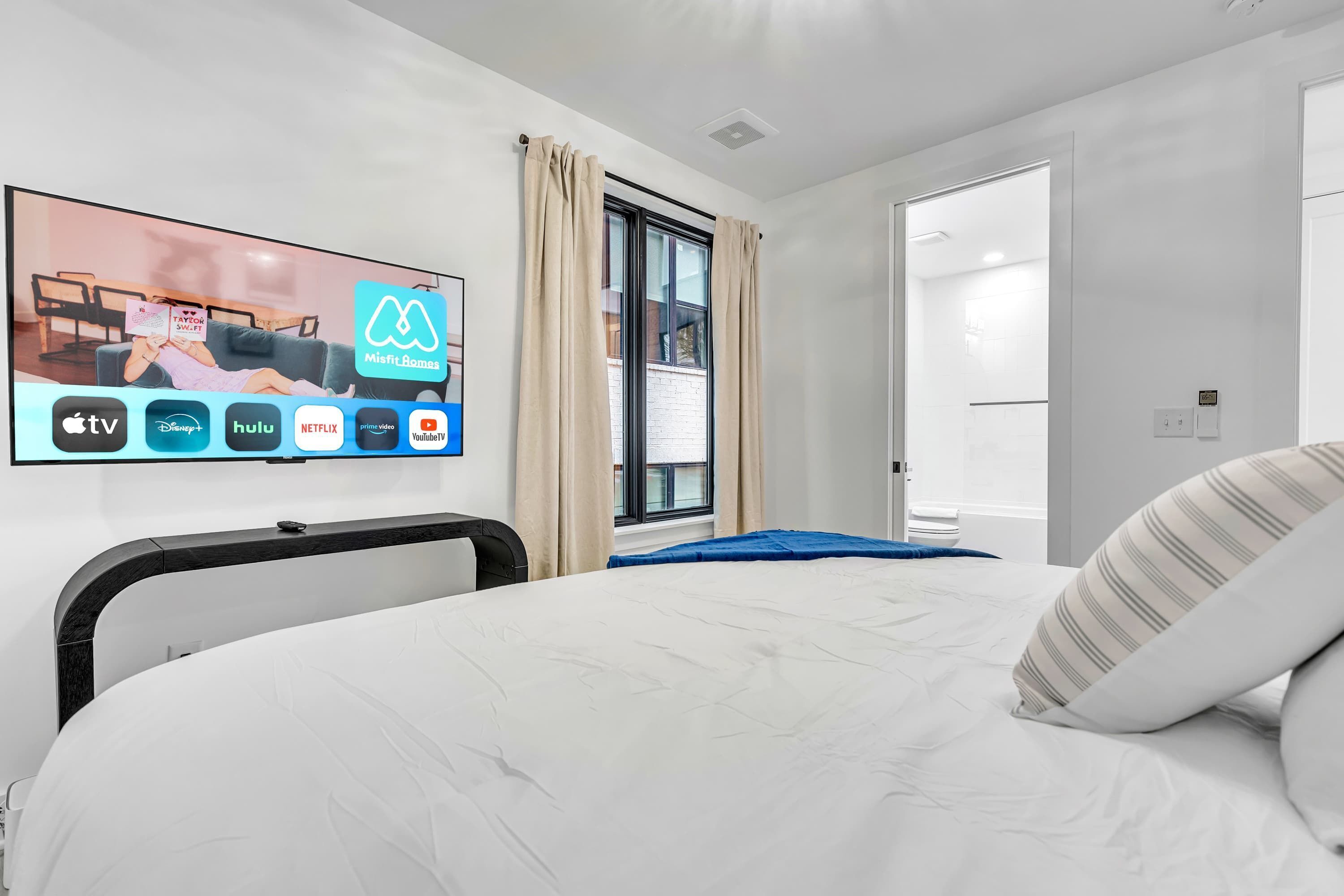Een moderne hotelkamer met een grote flatscreen televisie waarop streamingdiensten worden weergegeven, een opgemaakt bed met een blauwe deken, en uitzicht buiten door een raam met gordijnen.