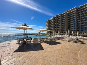 Área de piscina em resort com espreguiçadeiras e guarda-sóis, um edifício grande ao fundo e céu claro.