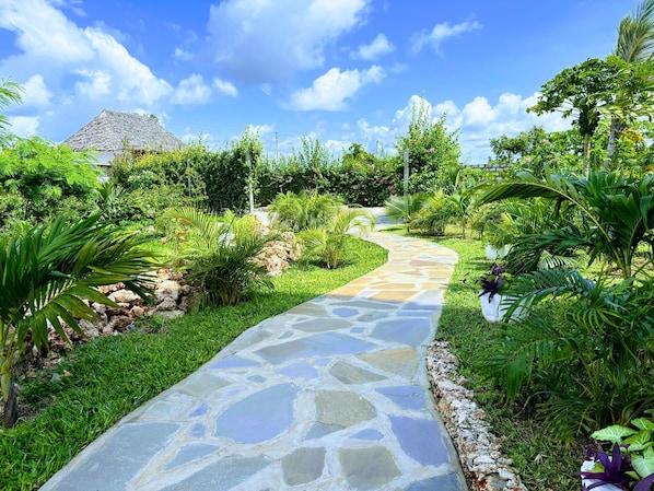 A stone pathway leading through a lush garden with tropical plants and a thatched-roof structure in the background.