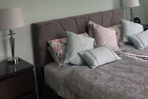 Ein ordentlich bezogenes Bett mit einem gemusterten Überwurf und mehreren Kissen, ein Nachttisch mit Lampe sowie eine Kommode mit Schubladen.