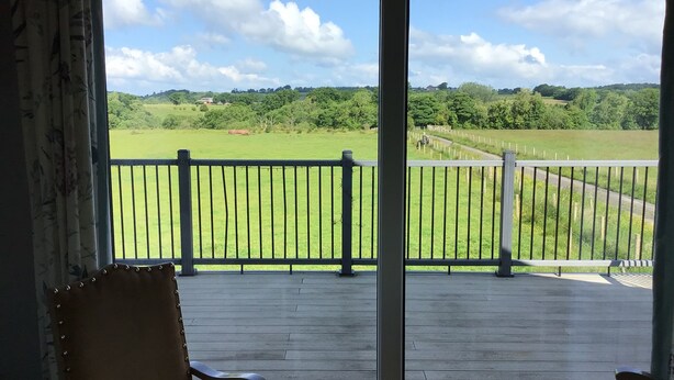 A view from a room with a wooden deck and a metal railing, overlooking a green field and a road with a fence. - More Than Just a Room Apartment Carlisle / Borders (Carlisle)