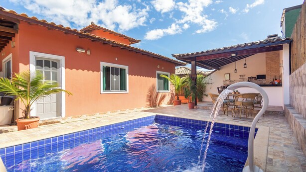 Uma piscina com um elemento aquático, um pátio com mobiliário de exterior e um edifício com telhado em azulejos.