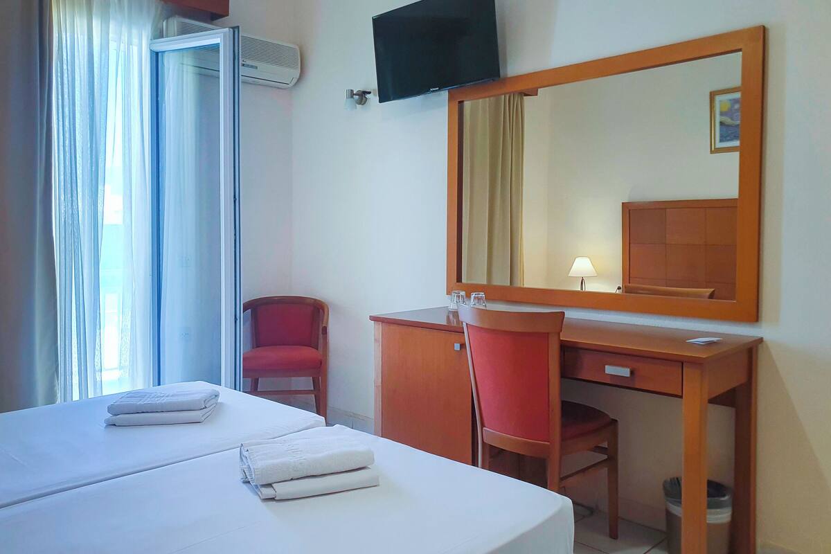 Una habitación de hotel con cama, escritorio, silla, televisor y ventana con cortinas.