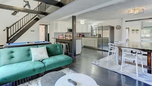 Un espacio habitable amplio con un sofá verde, una mesa de comedor con sillas, una cocina al fondo y una escalera que sube a un nivel superior.