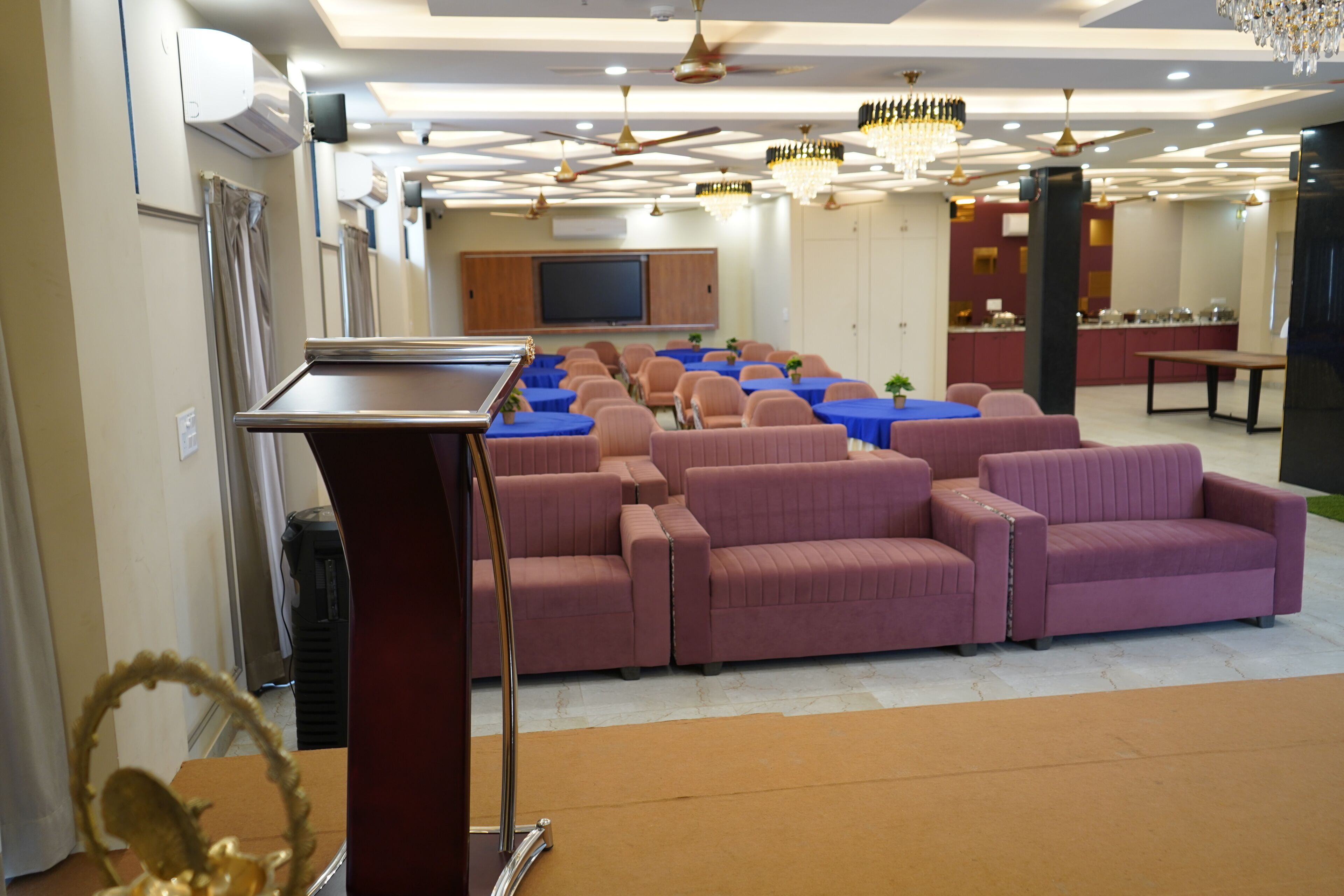 Un salón de conferencias con un atril, filas de sofás, mesas y una pantalla de televisión.
