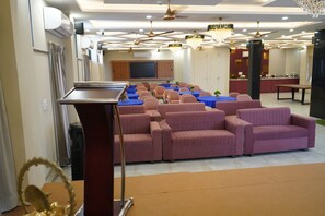 Un salón de conferencias con un atril, filas de sofás, mesas y una pantalla de televisión.