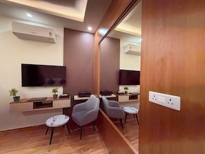 A modern hotel room with a flat-screen TV, wooden flooring, a desk with a chair, and a small round table. - Aanchal Palace Hotel  (Jaipur)