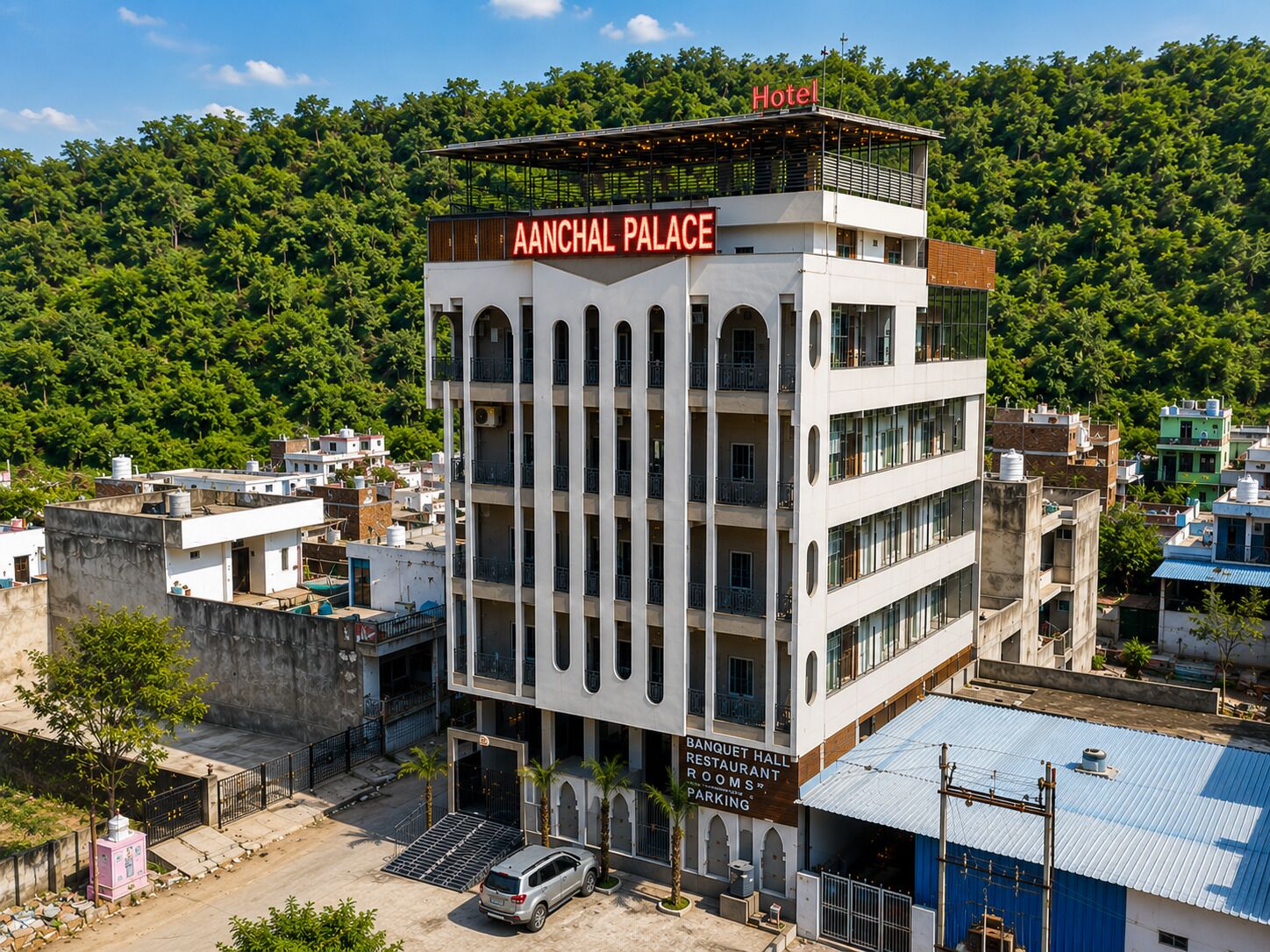 Un hotel de varios pisos llamado 'AANCHAL PALACE' con un letrero que indica 'SALÓN DE FIESTAS, RESTAURANTE, HABITACIONES' y 'ESTACIONAMIENTO'.