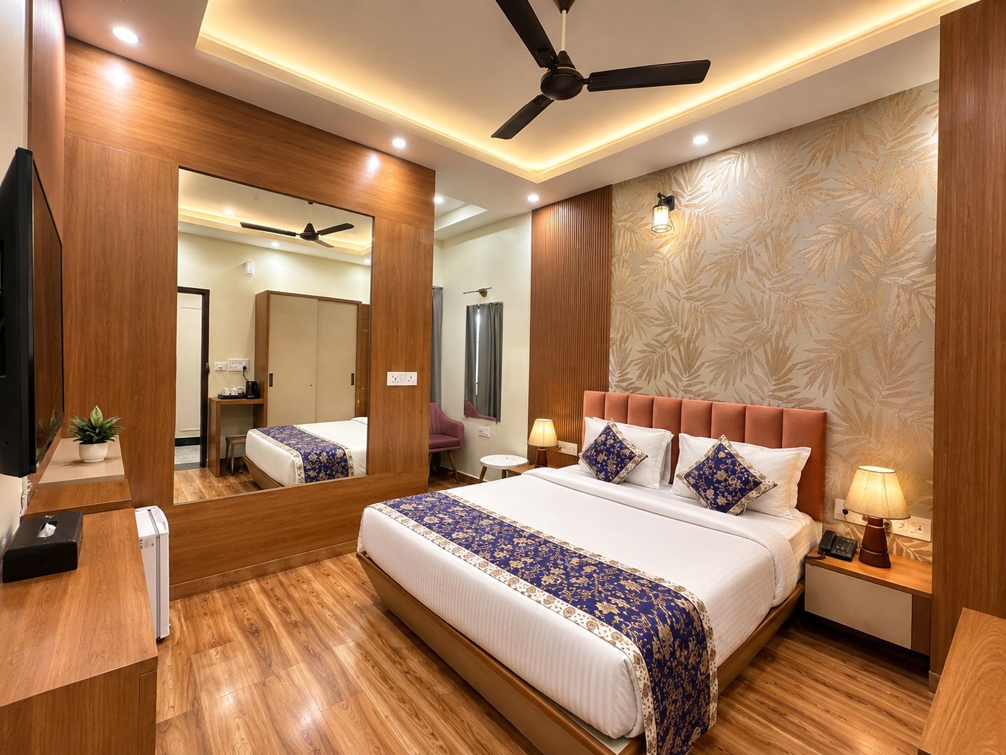 A hotel room with a large bed, a ceiling fan, a mirror, a bedside table with a lamp, and a wooden floor.