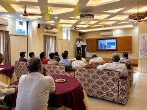 A conference room with a speaker at a podium, a screen displaying a presentation, and attendees seated at round tables. - Aanchal Palace Hotel  (Jaipur)