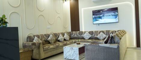 A modern living room with a sectional sofa, a smaller armchair, a marble coffee table, and a flat-screen TV mounted on the wall.