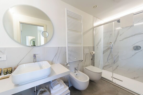 Un baño moderno con un espejo ovalado grande, un lavamanos blanco, un bidé, un inodoro y una ducha con mampara de vidrio.