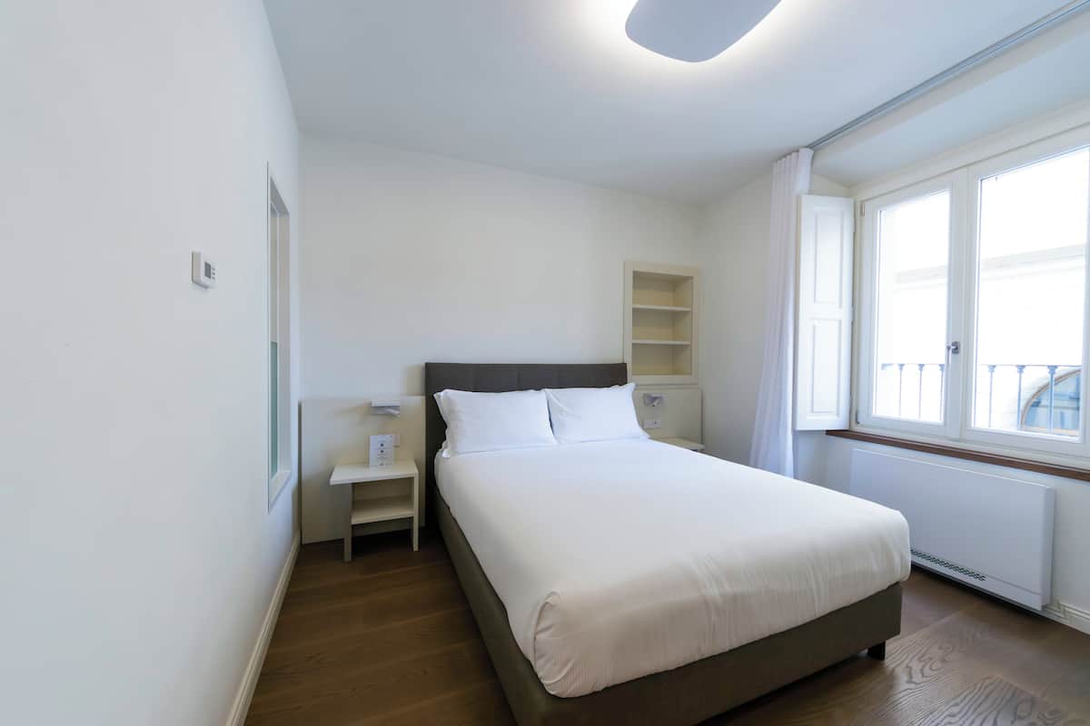 Quarto moderno com cama grande, mesa de cabeceira, prateleira embutida e janela com cortinas.